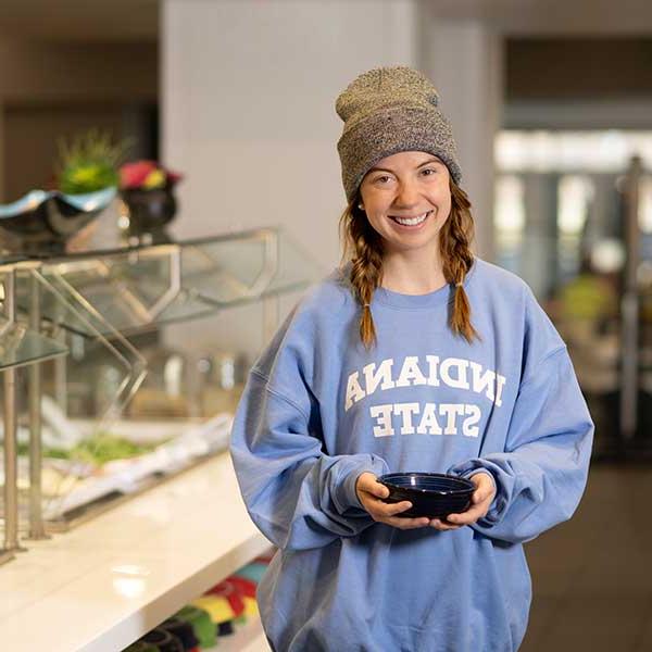 A smiling female student in a grey wool hat and a blue Indiana State sweatshirt holds a bowl next to the salad bar in the dining hall.