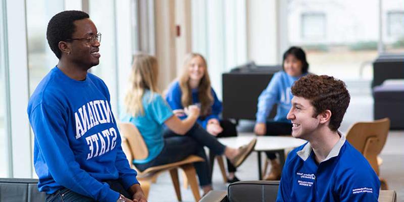 Two students wearing blue Indiana State shirts laugh together in the Commons areas of a residence hall. Several other students are seen sitting and smiling in the background.