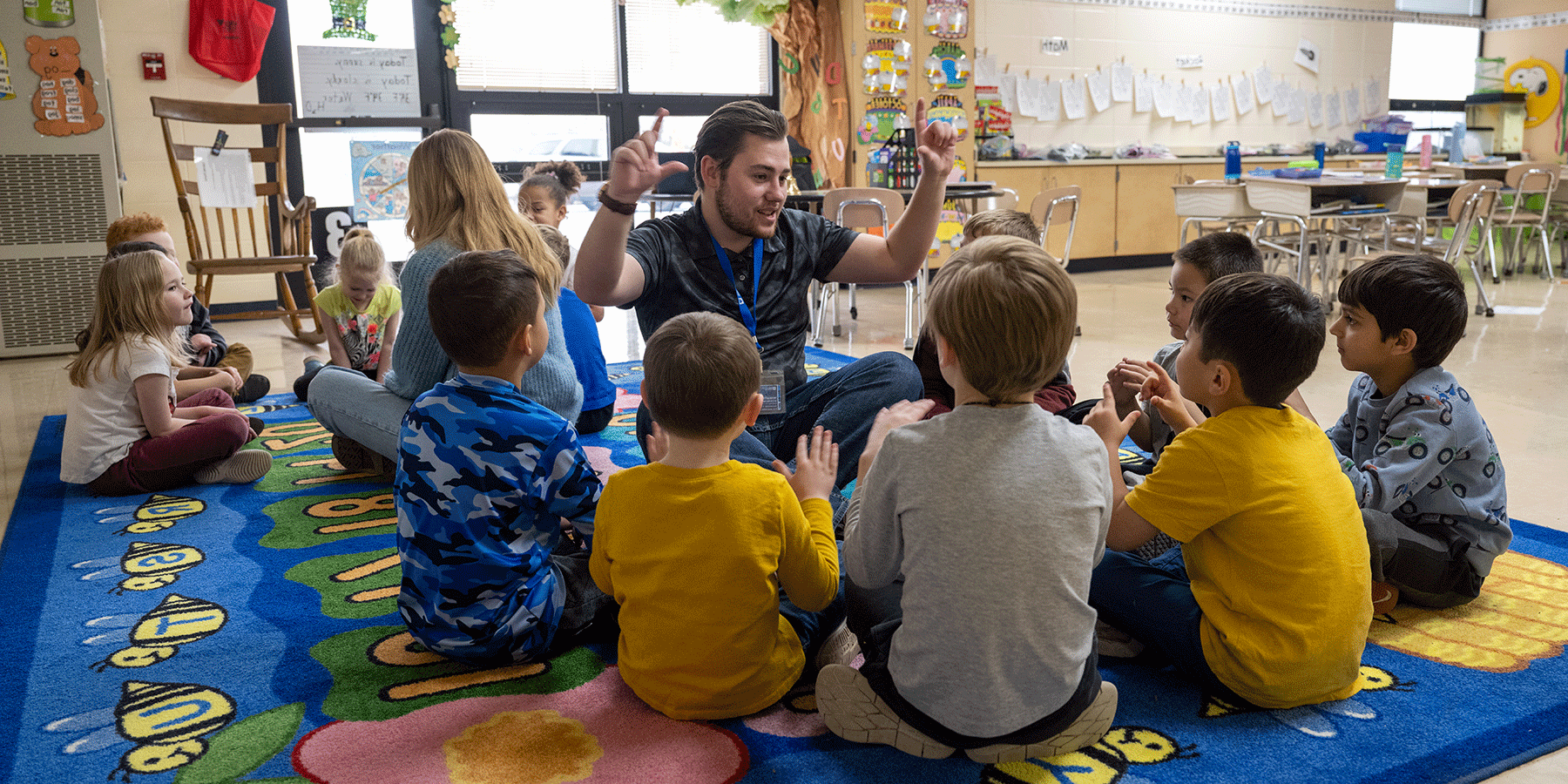 A classroom with 13 young children of mixed races, wearing a variety of yellow, blue, and grey clothing. The children are divided into two groups, sitting in circles on a blue, green, and yellow-colored floor rug. In the left group, a white male sits with the students. He has short brown hair with a matching beard. He wears a black T-shirt and blue jeans. He is holding up both hands as he looks at the students. In the right group, a white female sits with the students.