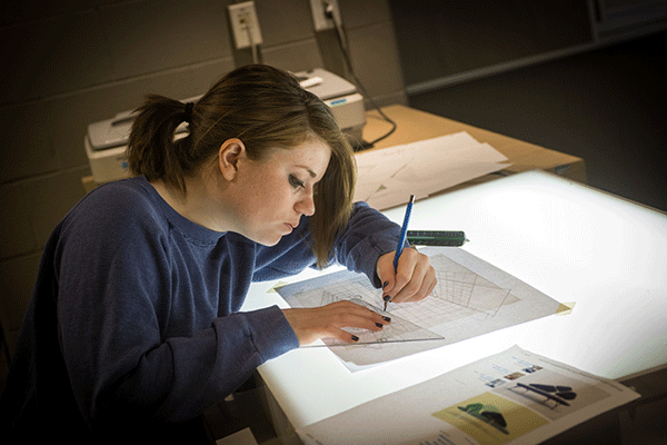 In a lab space, a white female student with brown hair pulled back into a ponytail, sits leaned over an illuminated drafting table. She is wearing a blue long-sleeved shirt. She holds a pencil in her right hand, a protractor in her left hand, and she is drawing a design on a page.