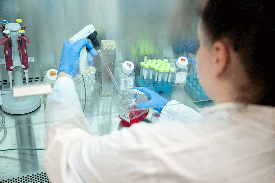 The lab tech faces away from the camera while using lab equipment to sample a substance in a container.