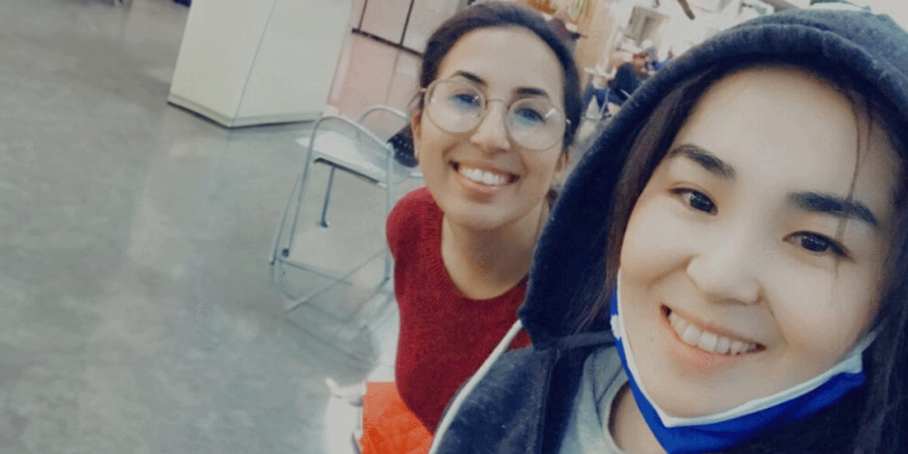 Two female students of Asian heritage pose for a selfie. On the left is a female student with brown hair. She wears a dark grey jacket with the hood up, and a blue mask pulled down from her mouth. On the right is a female student with brown hair pulled back. She wears glasses and a red sweater. Behind them are cabinets, a chair, and other individuals blurred in the background.