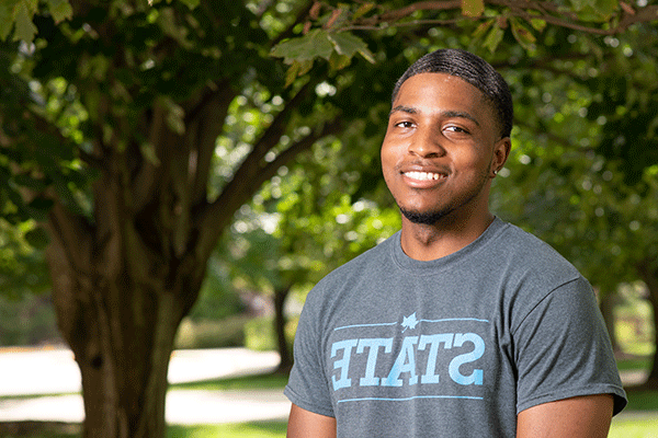 A smiling Black male student with black hair stands outside on campus with green, shady trees and sunshine in the background. He wears a grey shirt with STATE on it in light blue.