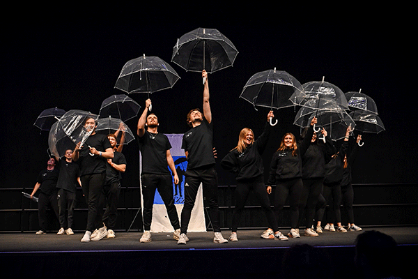 A group of male and female students dressed in black stand illuminated on a dark stage as they raise clear plastic umbrellas above their heads. A blue-and-white banner is visible behind them. One man on the right side holds his umbrella at a left angle to the side and not above his head like the others.