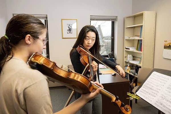 An Asian female professor with black shirt plays a violin while looking at sheet music as a White female student follows along.