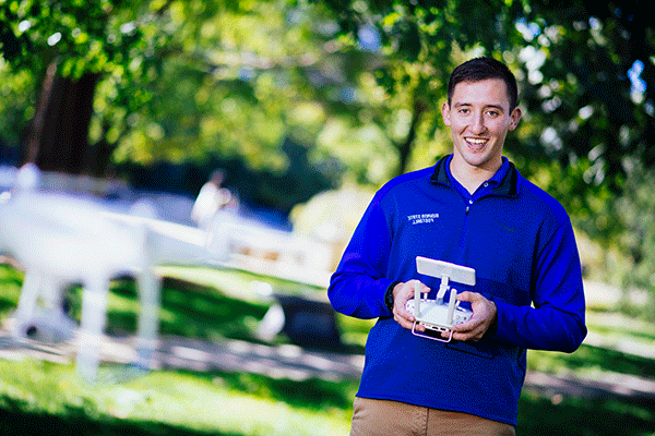 A white male student with short brown hair poses outside, wearing khaki pants and a blue zipped-up jacket with “Indiana State University” in white lettering in the top right corner of the jacket. He holds a white drone remote controller with a screen. A white drone is flying away from him, blurred in the photo. Green trees are visible in the background, also blurred in the photo. 