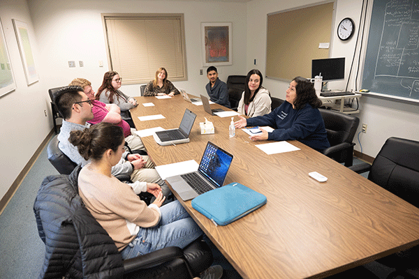 In a conference room, there are eight individuals of mixed races sitting at a table. They are wearing a variety of grey, white, blue, and pink clothing. Laptops are on the brown conference table. 