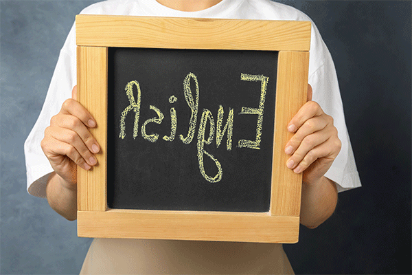 An individual in a white shirt, whose hands are visible and whose face is not, holds a square slate chalkboard framed in wood. The word ‘English’ is written on the chalkboard in large yellow letters.  